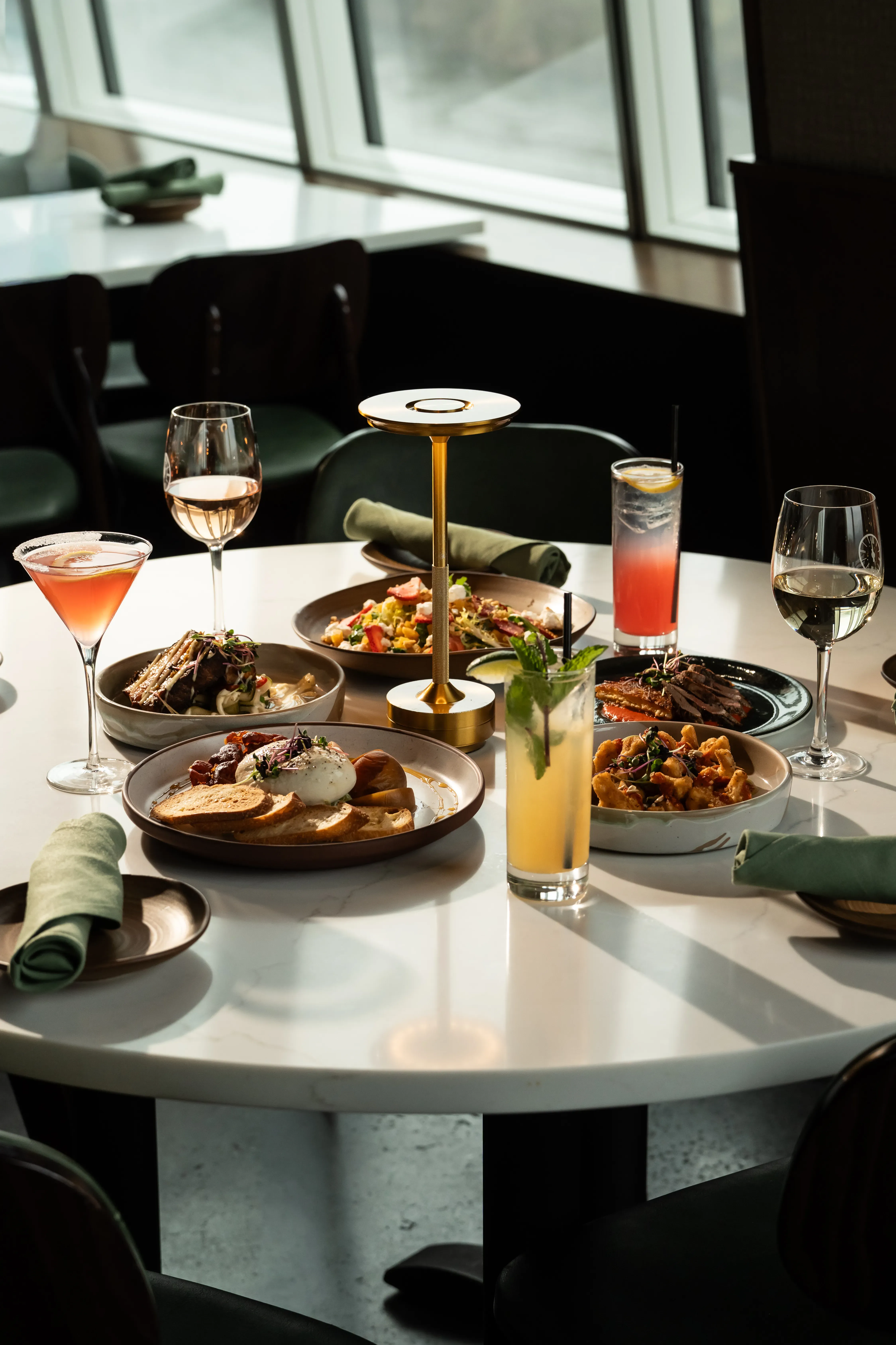 A round marble table at Thyme set with cocktails, wine, burrata toast, salad, sliced steak, and other small plates, lit by a brass table lamp and warm window light.