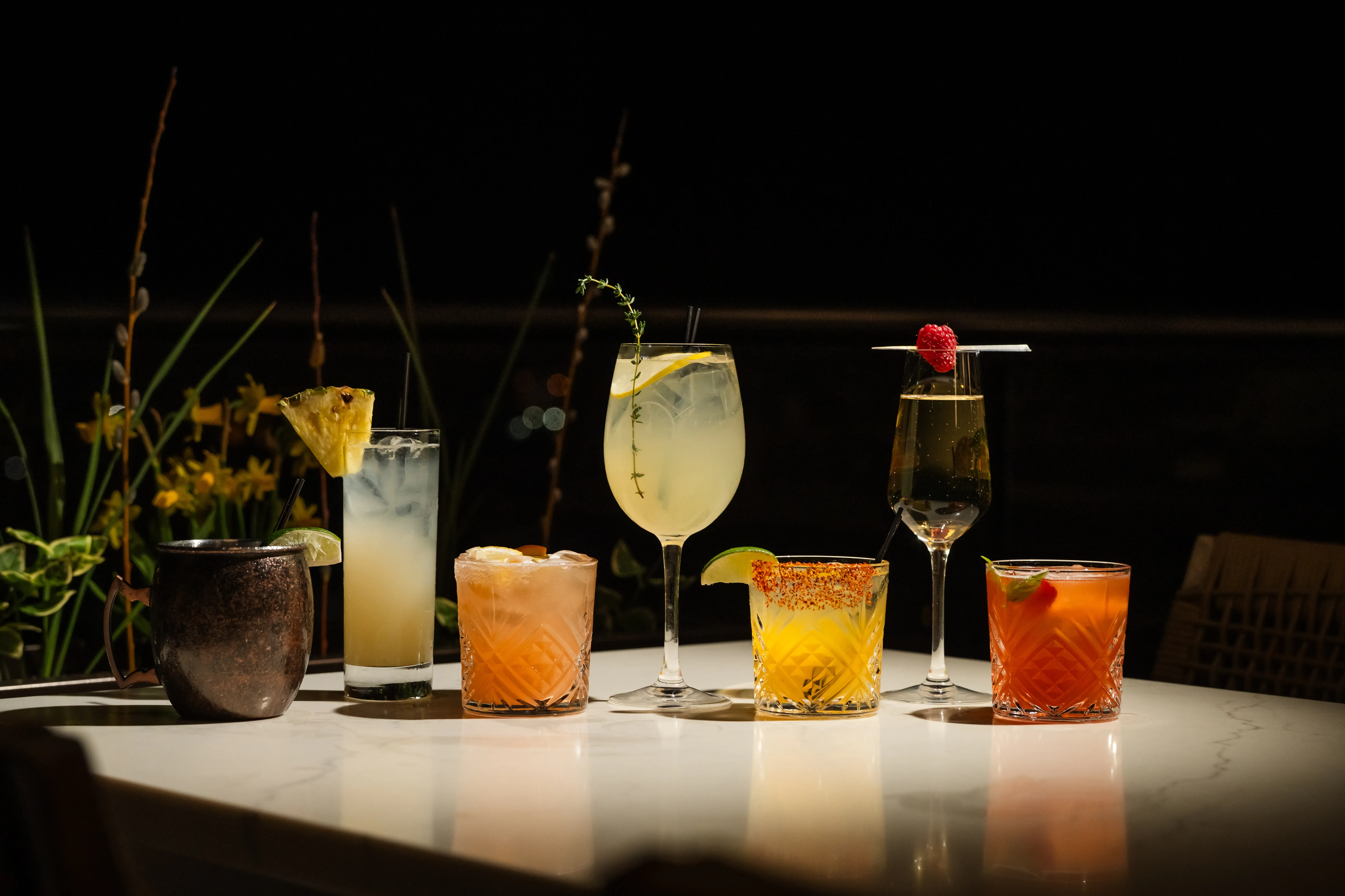 A lineup of seven Thyme cocktails on a marble bartop, including a copper mug mule, a thyme-and-citrus highball, a pineapple-garnished sour, a champagne flute with a raspberry, and citrus-rimmed rocks drinks, set against a moody dark backdrop.
